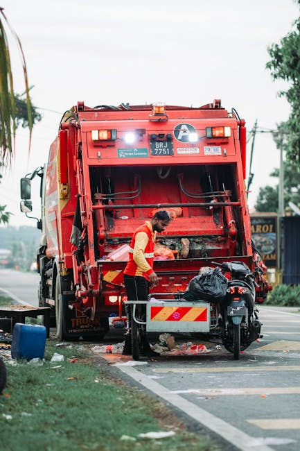 A waste collection worker wearing a red and beige uniform stands at the rear of a large red rubbish collection truck, which is parked on the side of a street. The back of the truck is open, showing compartment details and hydraulic mechanisms. The worker appears to be detaching or inspecting the waste hopper, with a black garbage bag placed nearby on the ground. The scene includes a small waste bin or container on the pavement, with some scattered debris and trash around the working area. In the background, there are trees, utility poles, and a building with signage, indicating an urban or suburban environment. The sky is overcast, providing diffuse natural lighting that highlights the textures of the truck's metal surface, the plastic trash bags, and the pavement. This image illustrates an example of independent waste collection or rubbish removal service, emphasizing practical methods of rubbish disposal outside of municipal systems.