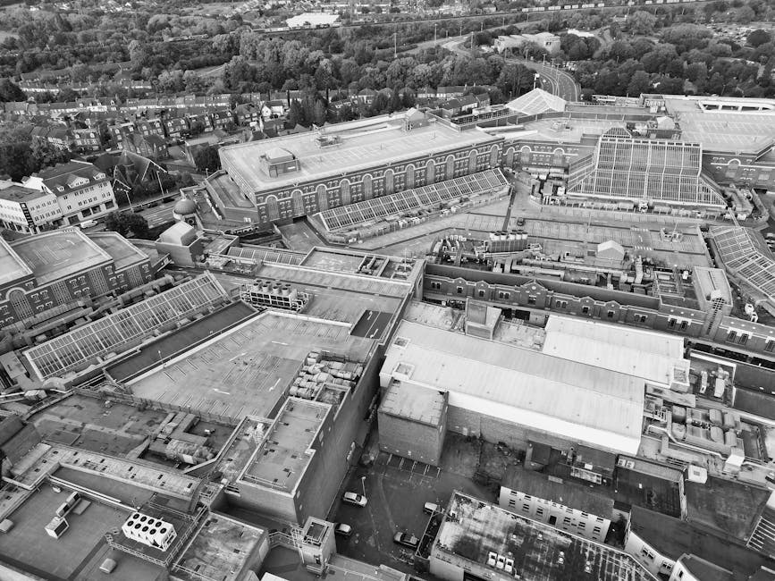 Aerial view of a large industrial and commercial complex featuring numerous interconnected flat roofs with various ventilation units, skylights, and roofing materials. The rooftops include metal and glass structures, with some sections showing clear glass panels that allow natural light into the buildings below. Surrounding the main structures are parking areas with several parked vehicles and adjacent roads, with residential areas and green spaces in the background. The environment appears to be well-organized, with clear pathways and service access points. The image's lighting is natural, likely taken during daylight hours, emphasizing the textures of the roofs' materials. Visible equipment on the roofs includes HVAC units and ventilation pipes, contributing to an industrial aesthetic. The scene depicts a typical urban industrial zone, consistent with premises where waste management services such as rubbish removal might be needed, especially considering the proximity of various buildings and storage areas which could generate waste for disposal or collection by companies like Rubbish Collection Watford, known for alternative waste handling solutions.