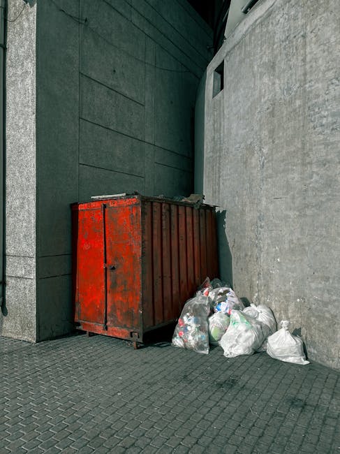 A waste collection worker wearing a red and beige uniform stands at the rear of a large red rubbish collection truck, which is parked on the side of a street. The back of the truck is open, showing compartment details and hydraulic mechanisms. The worker appears to be detaching or inspecting the waste hopper, with a black garbage bag placed nearby on the ground. The scene includes a small waste bin or container on the pavement, with some scattered debris and trash around the working area. In the background, there are trees, utility poles, and a building with signage, indicating an urban or suburban environment. The sky is overcast, providing diffuse natural lighting that highlights the textures of the truck's metal surface, the plastic trash bags, and the pavement. This image illustrates an example of independent waste collection or rubbish removal service, emphasizing practical methods of rubbish disposal outside of municipal systems.