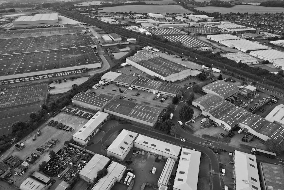 An aerial view of an industrial or commercial area showing multiple large warehouse and factory buildings with flat roofs mostly covered with roofing sheets, some with skylights or vents. The buildings are arranged in a grid pattern with parking lots adjacent, containing numerous vehicles including vans, cars, and trucks. A few smaller structures are visible alongside the main warehouses, and a network of roads and driveways connects all the areas. The landscape is flat with trees and green patches interspersed between buildings, and in the distance, there are additional warehouse units, possibly storage or distribution centers. The overall scene is captured in black and white, highlighting the textures of the roofing materials, the asphalt parking surfaces, and the density of the industrial site, presenting a clear overview suitable for understanding private waste or rubbish management operations in a typical business park environment.
