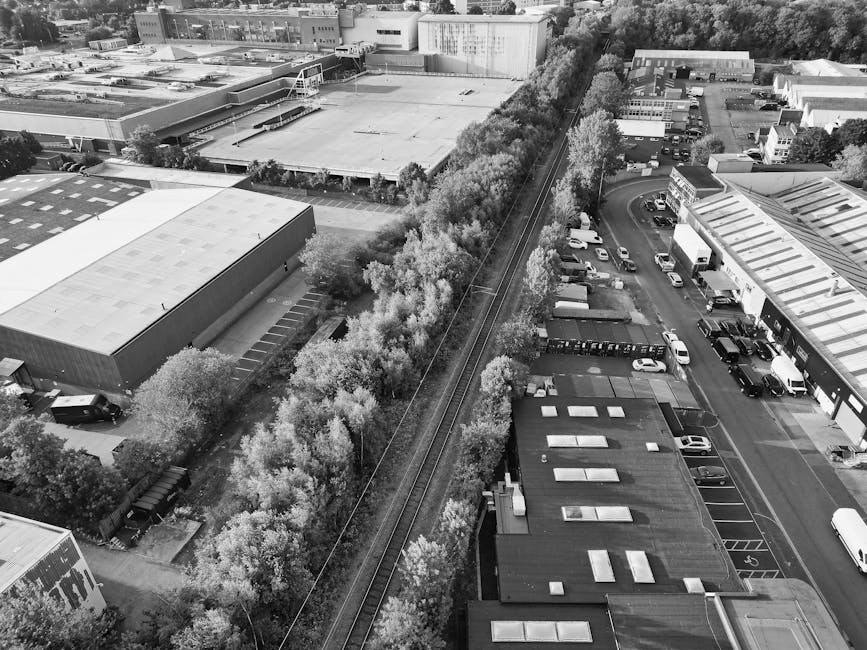 An aerial black-and-white photograph showing a long, narrow line of densely packed deciduous trees with light-colored leaves running parallel to a railway track that cuts diagonally across the image. To the left of the trees, there are industrial buildings and warehouses with flat roofs, some with visible vents or skylights, separated by small parking areas. To the right, a commercial area features a row of parked cars and trucks along a street, with some trucks spaced out and others clustered near buildings with flat roofs. The environment appears urban, with surrounding structures and streets, and the trees serve as a natural boundary between the industrial and commercial zones. The lighting is even, with soft shadows indicating an overcast sky or diffuse daylight, and the scene reflects a typical urban landscape where private waste collection services might operate to manage rubbish removal outside of local authority provisions, with Rubbish Collection Watford potentially involved in handling waste from nearby commercial or industrial properties.