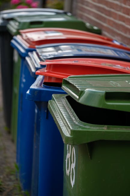 A row of several large wheeled waste bins arranged along a paved pathway beside a brick wall. The bins are primarily blue, with the nearest green bin in the foreground and others in blue, red, and green colors visible behind it. The lids of the bins are closed, with some lids slightly angled, and the bins appear to be made of durable plastic with textured surfaces and molded handles. The surrounding environment includes a brick wall on the right side and a blurred background suggesting an outdoor residential or communal area. The scene is lit with natural daylight, providing clear visibility of the detailed textures and colors of the bins, which are commonly used for private rubbish collection services, such as those offered by Rubbish Collection Watford for alternative waste handling or onsite clearance. Overall, the image depicts a typical waste disposal setup aligned with rubbish removal services focused on efficient waste segregation and collection.